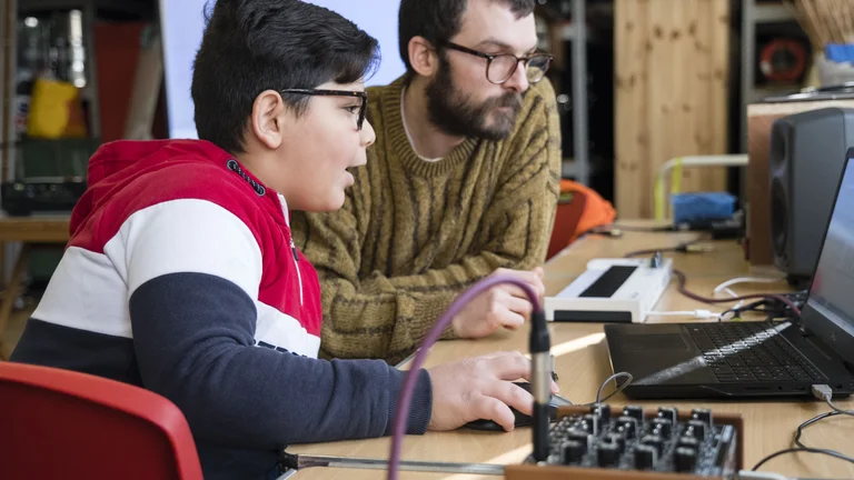 A music tutor and a young person looking at a laptop. The equipment around them is for electronic music production. 