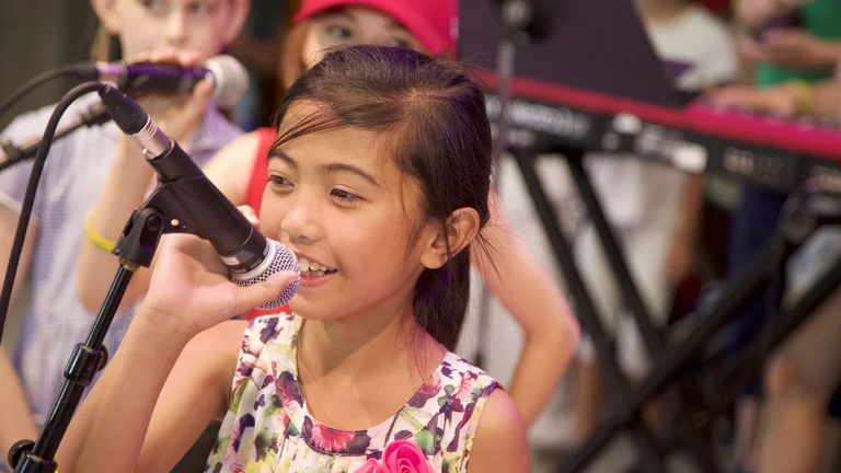 A child smiling and singing into a microphone. Two other children are standing behind them and a keyboard is in the background.