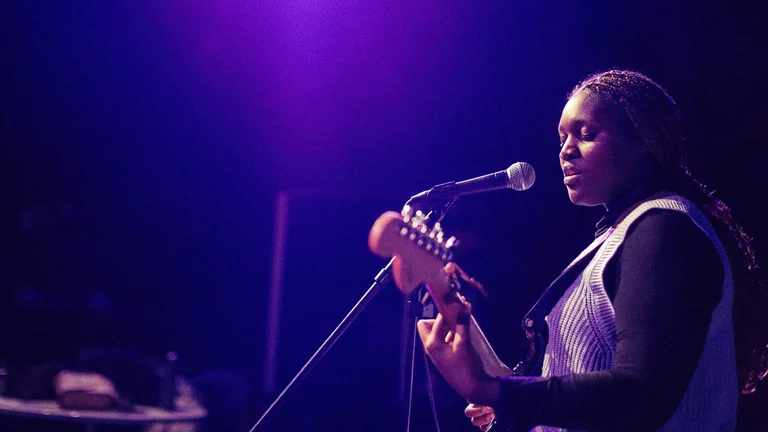 A woman singing and playing guitar, she is lit in a deep purple