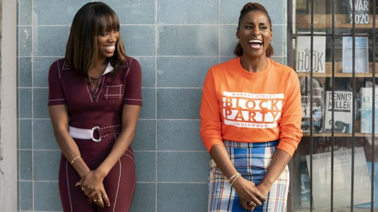 Two black people standing and laughing. One is wearing a burgundy dress, the other is wearing a orange jumper and patterned trousers.