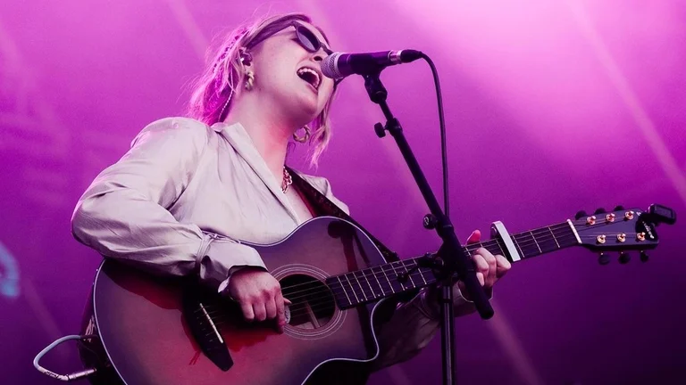 Young person singing on stage with a guitar, wearing sunglasses