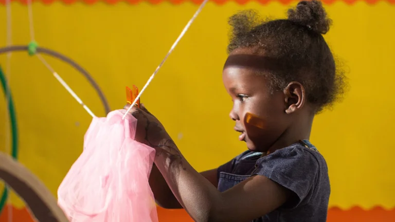 A child playing with a peg and fabric on a string line. The background is bright yellow.