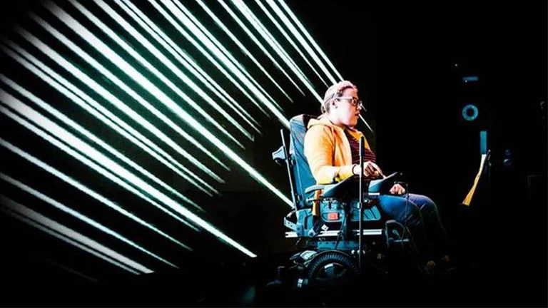 A woman on a stage sitting in an electric wheelchair, bright streaks of white light shine behind her