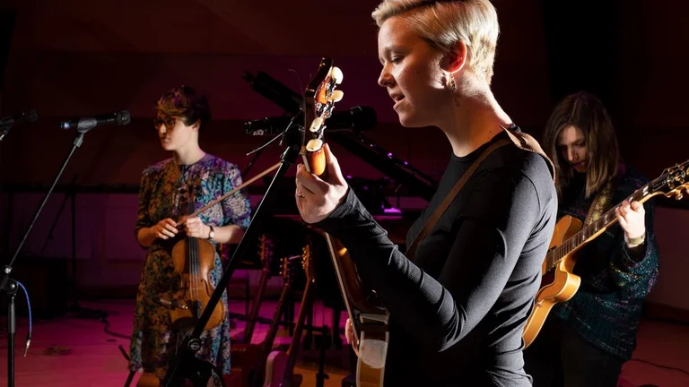 Person with short blond hair playing electric guitar on stage and singing, 2 other young people accompanying behind on guitars