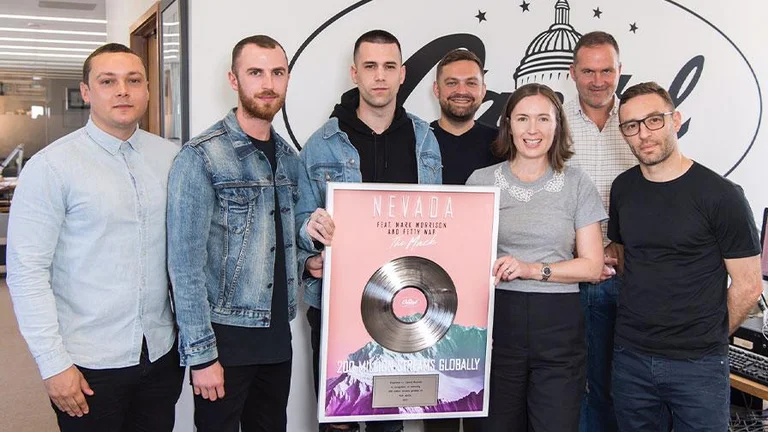 7 people standing in a row, some holding up a framed vinyl