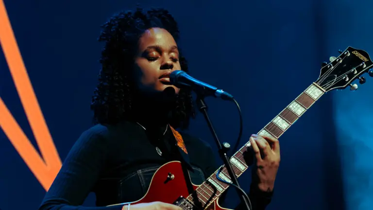 adjua closes her eyes as she sings into the mic and plays an electric guitar