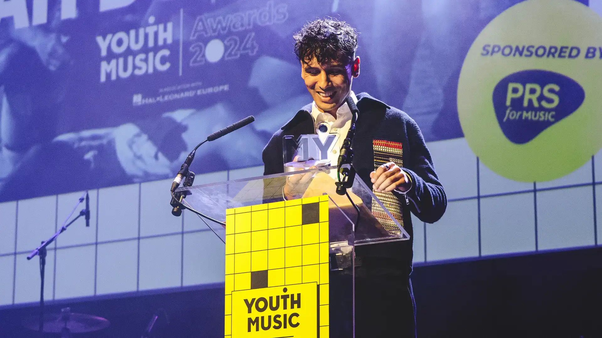 Noah Bouchard smiling at his award on stage at the YMA24, at a yellow lecturn