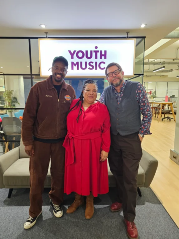 Matt the CEO of Youth Music standing with Guvna B and Charlotte Edgeworth in the Youth Music office in front of a Youth Music sign