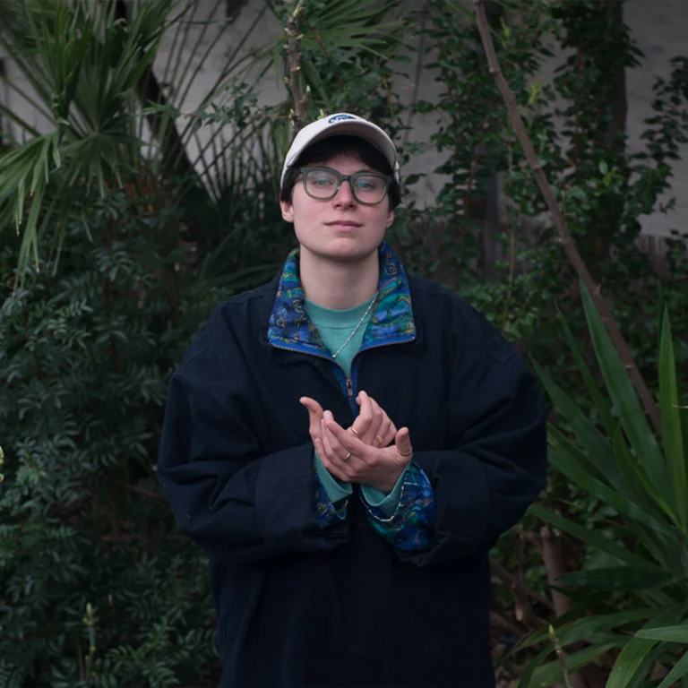 a person stands in front of some foliage and wears glasses and a white cap