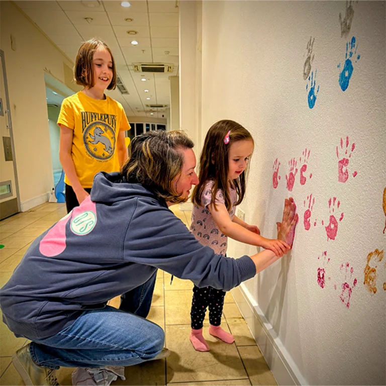 an adult helping two children paint handprints onto a white wall