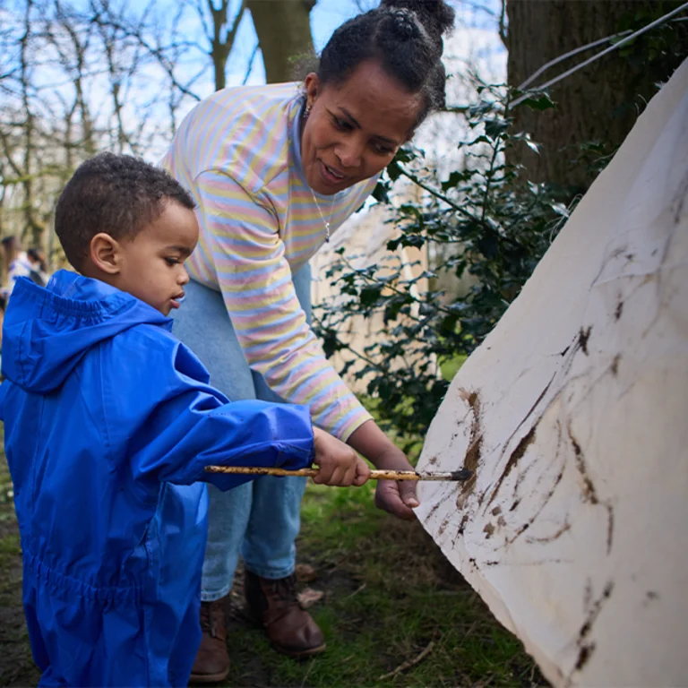 A young child in a blue raincoat painting outside in a wood with a smiling adult